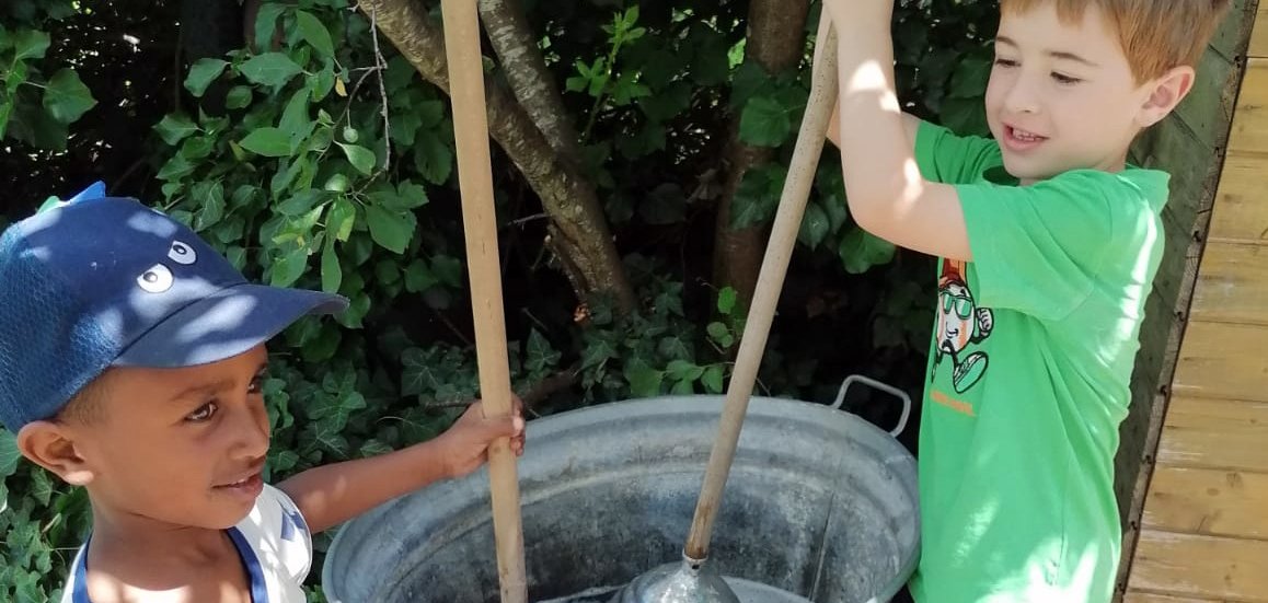 Auf dem Foto sind zwei Kinder zu sehen, die gemeinsam an einer historischen Wascheinrichtung arbeiten. Mit sogenannten Waschstampfern bewegen sie Wasser und Seifenlauge in einem großen Metallbottich, während vor ihnen ein traditionelles Waschbrett steht. Die Szene vermittelt spielerisch, wie aufwendig die Wäschepflege früherer Generationen war, und lädt dazu ein, historische Alltagsarbeit selbst auszuprobieren.