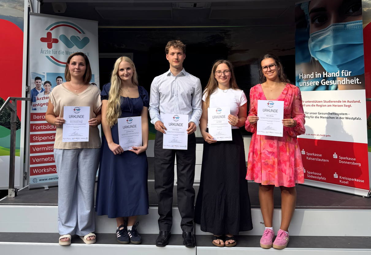ÄfdW Stipendiaten 2026 Auf dem Foto stehen fünf junge Stipendiatinnen und Stipendiaten nebeneinander auf einer Bühne und halten jeweils ihre Urkunde in den Händen. Hinter ihnen befinden sich Roll-ups des Vereins „Ärzte für die Westpfalz e. V.“ mit Informationen zum Stipendienprogramm und zur Förderung des medizinischen Nachwuchses. Die Aufnahme dokumentiert die feierliche Ehrung der Stipendiaten und vermittelt einen positiven, offiziellen Anlass.
