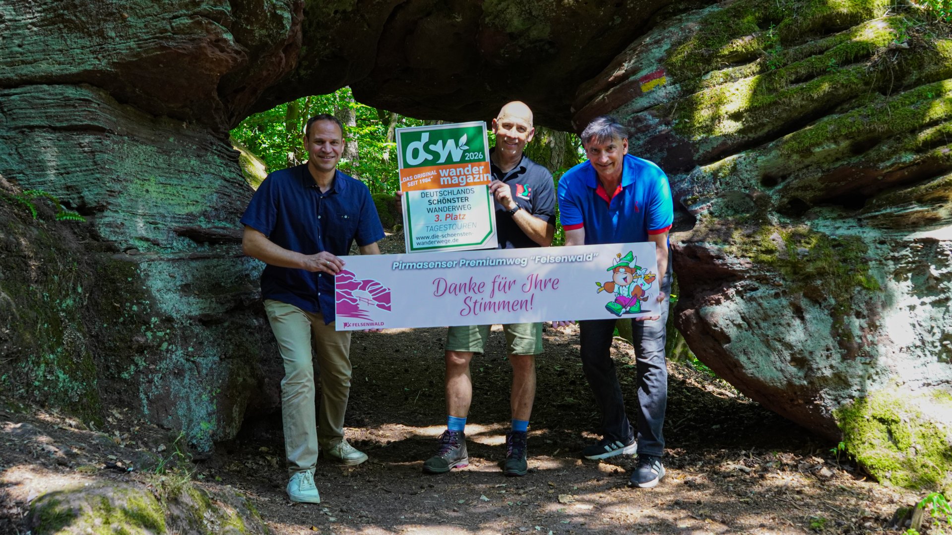 Felsenwald Auf dem Foto stehen drei Personen in einer bewaldeten Felsformation, die wie ein natürlicher Torbogen wirkt. Sie halten Schilder zum Premiumwanderweg „Felsenwald“ in Pirmasens. Ein Schild verweist auf die Auszeichnung als drittplatzierter Wanderweg bei einer Wahl zu Deutschlands schönsten Tagestouren, das andere bedankt sich mit der Aufschrift „Danke für Ihre Stimmen!“. Die Szene vermittelt Naturerlebnis, Wanderkultur und die Freude über die erzielte Platzierung.