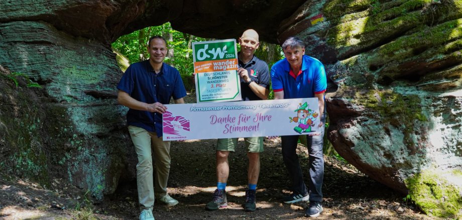 Felsenwald Auf dem Foto stehen drei Personen in einer bewaldeten Felsformation, die wie ein natürlicher Torbogen wirkt. Sie halten Schilder zum Premiumwanderweg „Felsenwald“ in Pirmasens. Ein Schild verweist auf die Auszeichnung als drittplatzierter Wanderweg bei einer Wahl zu Deutschlands schönsten Tagestouren, das andere bedankt sich mit der Aufschrift „Danke für Ihre Stimmen!“. Die Szene vermittelt Naturerlebnis, Wanderkultur und die Freude über die erzielte Platzierung.