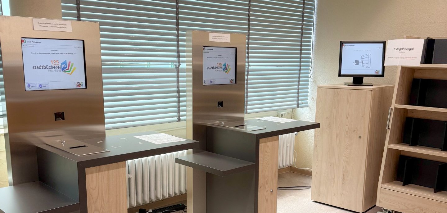 Selbstbucherterminals in der Stadtbücherei Pirmasens Auf dem Foto sind zwei moderne Selbstverbuchungsterminals in der Stadtbücherei zu sehen. Die Geräte verfügen über Touchscreens und ermöglichen Besucherinnen und Besuchern die eigenständige Ausleihe von Medien. Rechts im Bild befindet sich zudem ein Rückgabesystem mit Bildschirm und Regal zur automatisierten Medienrückgabe. Die Einrichtung wirkt hell, aufgeräumt und technisch modern. Die Aufnahme verdeutlicht die fortschreitende Digitalisierung der Stadtbücherei und die neuen Serviceangebote für Nutzerinnen und Nutzer.