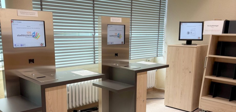 Selbstbucherterminals in der Stadtbücherei Pirmasens Auf dem Foto sind zwei moderne Selbstverbuchungsterminals in der Stadtbücherei zu sehen. Die Geräte verfügen über Touchscreens und ermöglichen Besucherinnen und Besuchern die eigenständige Ausleihe von Medien. Rechts im Bild befindet sich zudem ein Rückgabesystem mit Bildschirm und Regal zur automatisierten Medienrückgabe. Die Einrichtung wirkt hell, aufgeräumt und technisch modern. Die Aufnahme verdeutlicht die fortschreitende Digitalisierung der Stadtbücherei und die neuen Serviceangebote für Nutzerinnen und Nutzer.