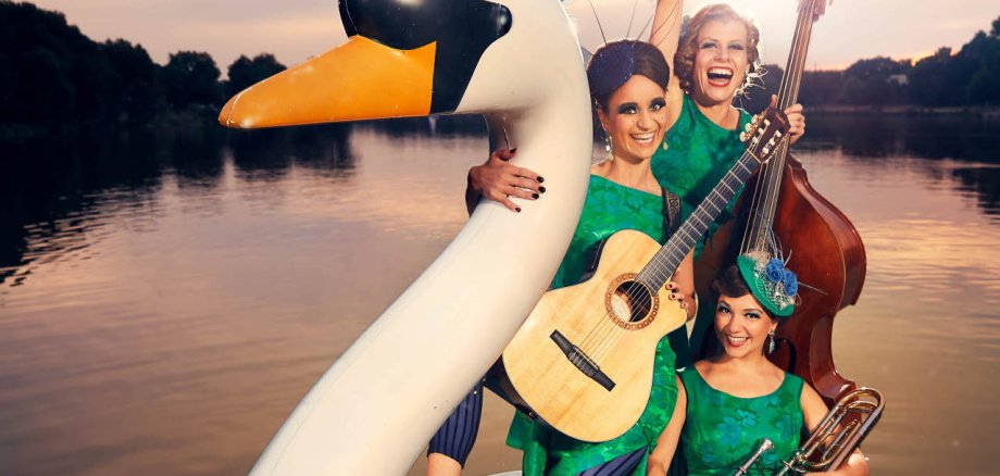 Zucchini-Sistaz Drei Musikerinnen in leuchtend grünen Retro-Kleidern posieren auf einem großen weißen Schwanen-Tretboot auf einem See. Eine Frau hält eine Gitarre, eine weitere steht mit einem Kontrabass hinter ihr und hebt lachend einen Arm in die Luft, während die dritte mit einer Posaune im vorderen Bereich des Bootes sitzt. Die Frauen tragen auffällige Kopfschmuck-Accessoires und präsentieren sich gut gelaunt im Stil einer nostalgischen Bühnenshow. Im Hintergrund ist die untergehende Sonne über dem ruhigen See zu sehen.