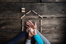 Familie Aufeinander gelegte Hände einer vierköpfigen Familie von oben fotografiert, obere Hand hält Herz und außenherum ein Haus mit Bauklötzen abgebildet
